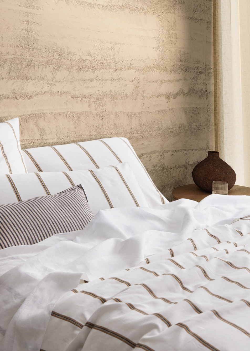 Close up of a white and neutral tone bed in front of a concrete wall. A vase sits on a bedside table.