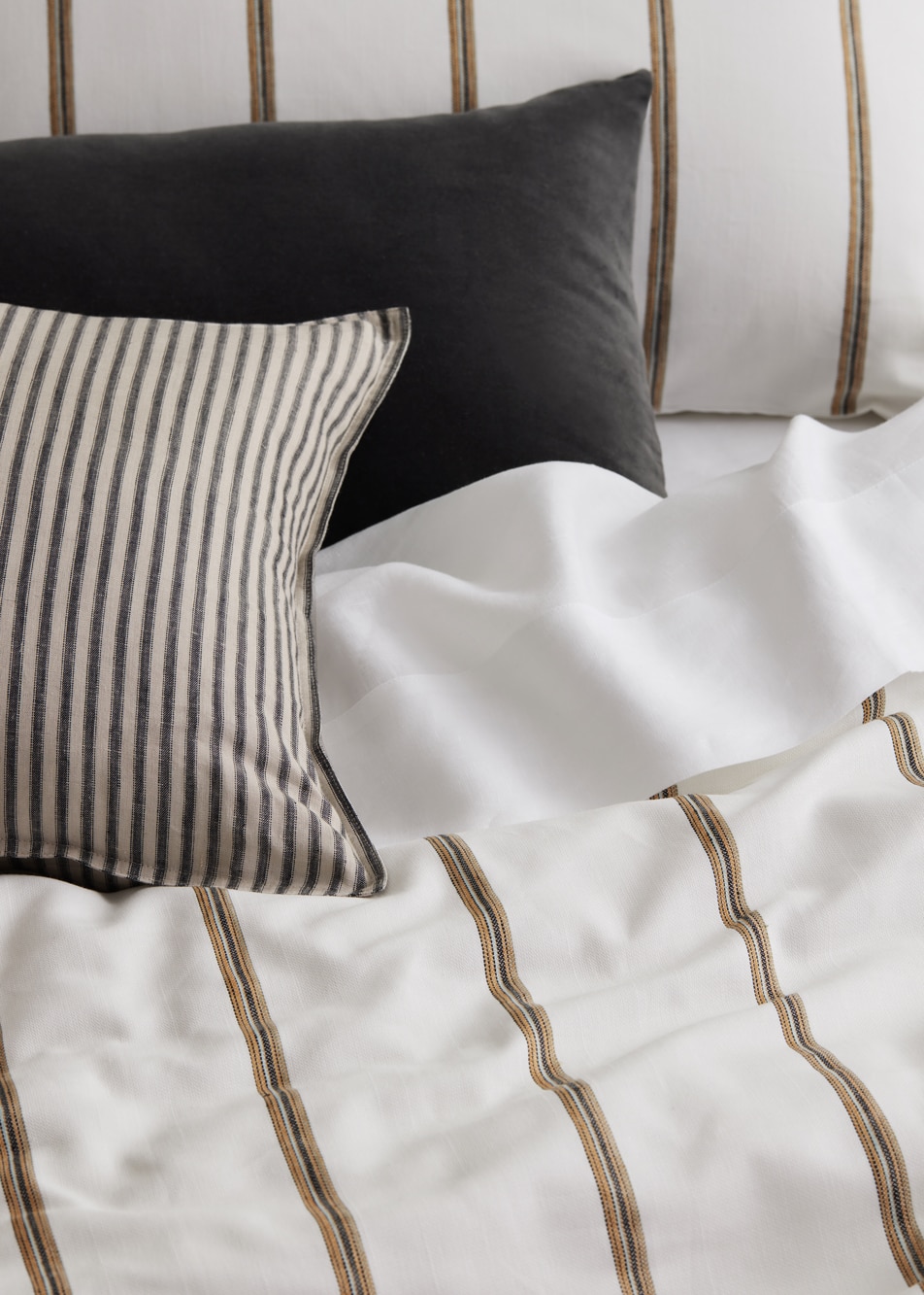 Close up of a white and neutral bed with one black cushion and one grey striped cushion on top